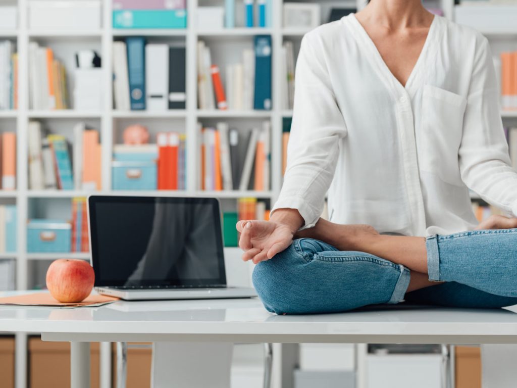 Woman in white blouse and jeans sitting on top of table in library, with computer and apple next to her.
