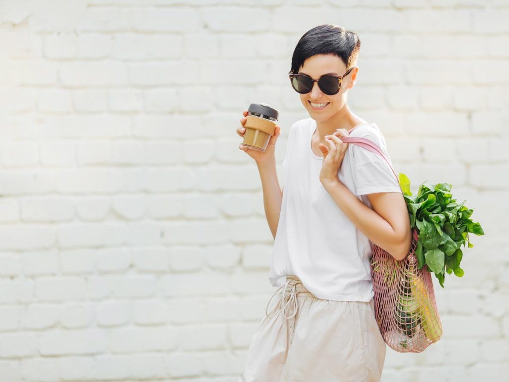 Woman holding zero waste coffee cup and pink bag filled with produce in front of white wall.