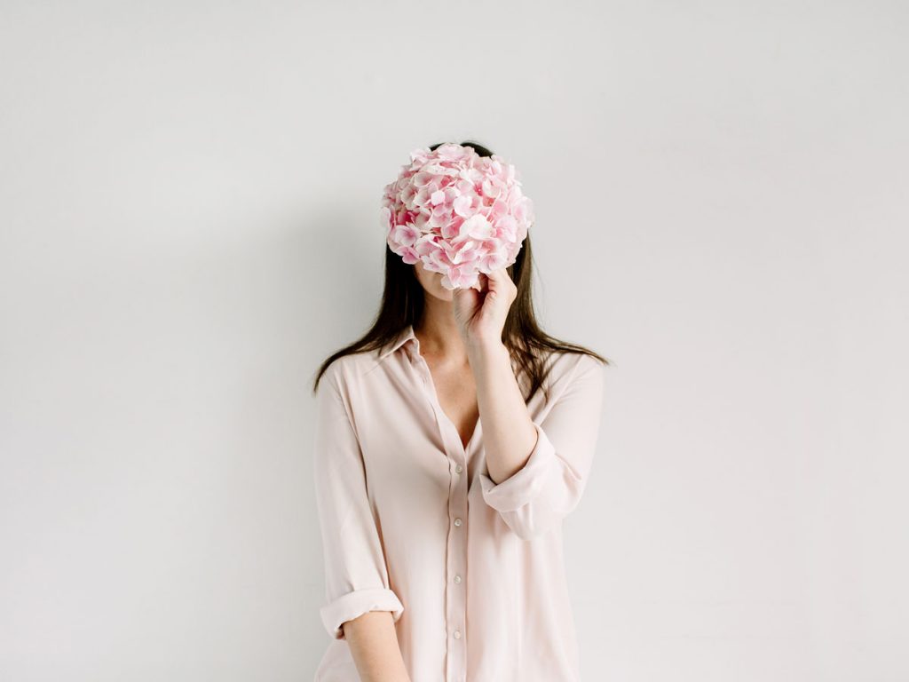 Woman in pink button down shirt holding pink flowers in front of face.