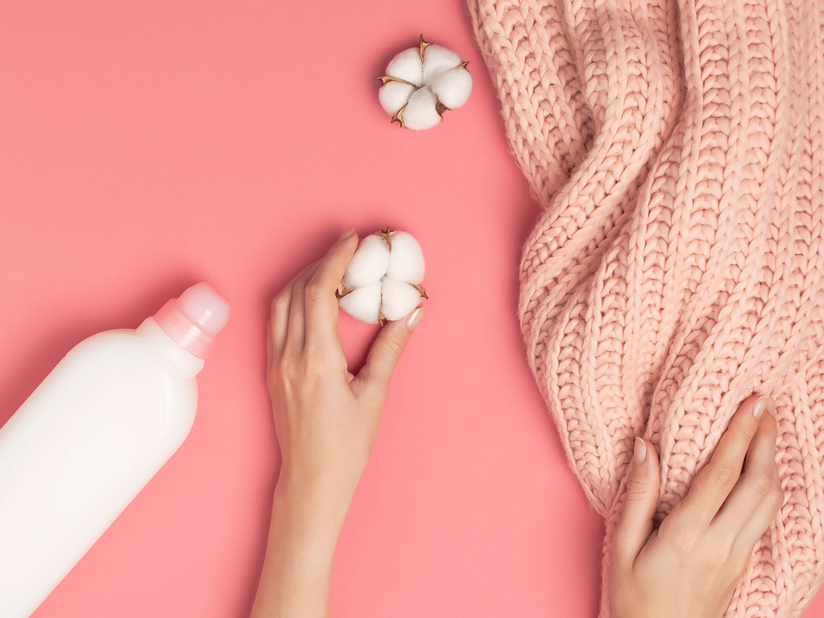 Flat lay of eco friendly fabric softener bottle, cotton fluff, and hands holding sweater.