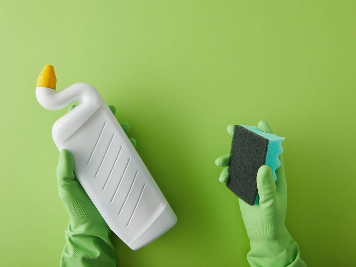 Gloved hands holding eco friendly toilet bowl cleaner and sponge against green background.