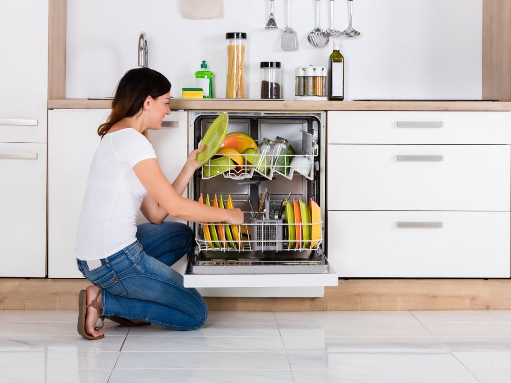 Woman in kitchen loading colorful plates and eco friendly dishwasher detergent into machine.